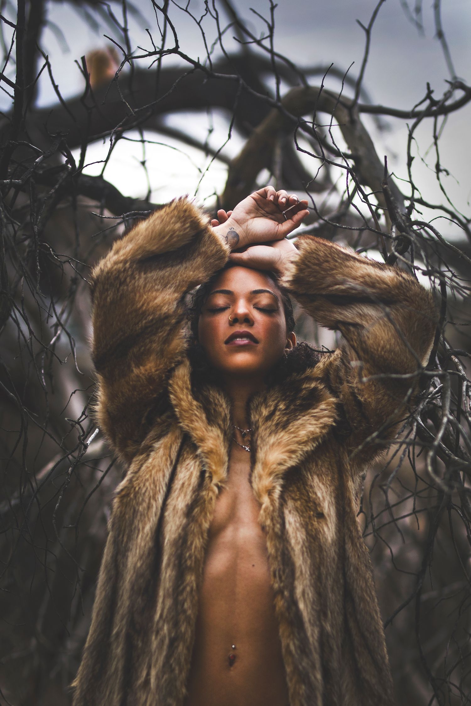 Woman in fur coat poses dramatically among bare branches in moody outdoor fashion photography.