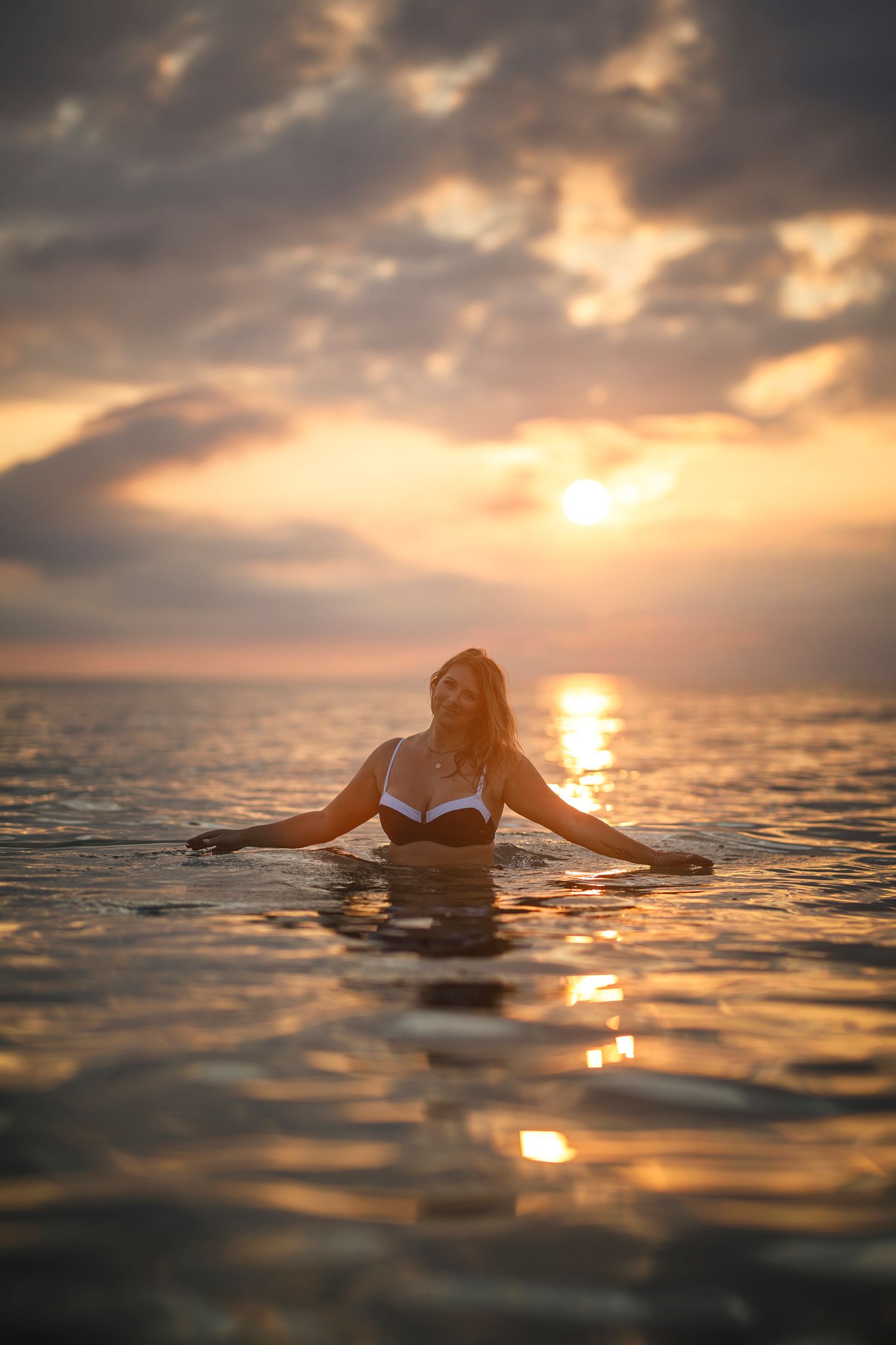 Woman in white bikini standing in ocean water during golden sunset with dramatic cloudy sky backdrop.