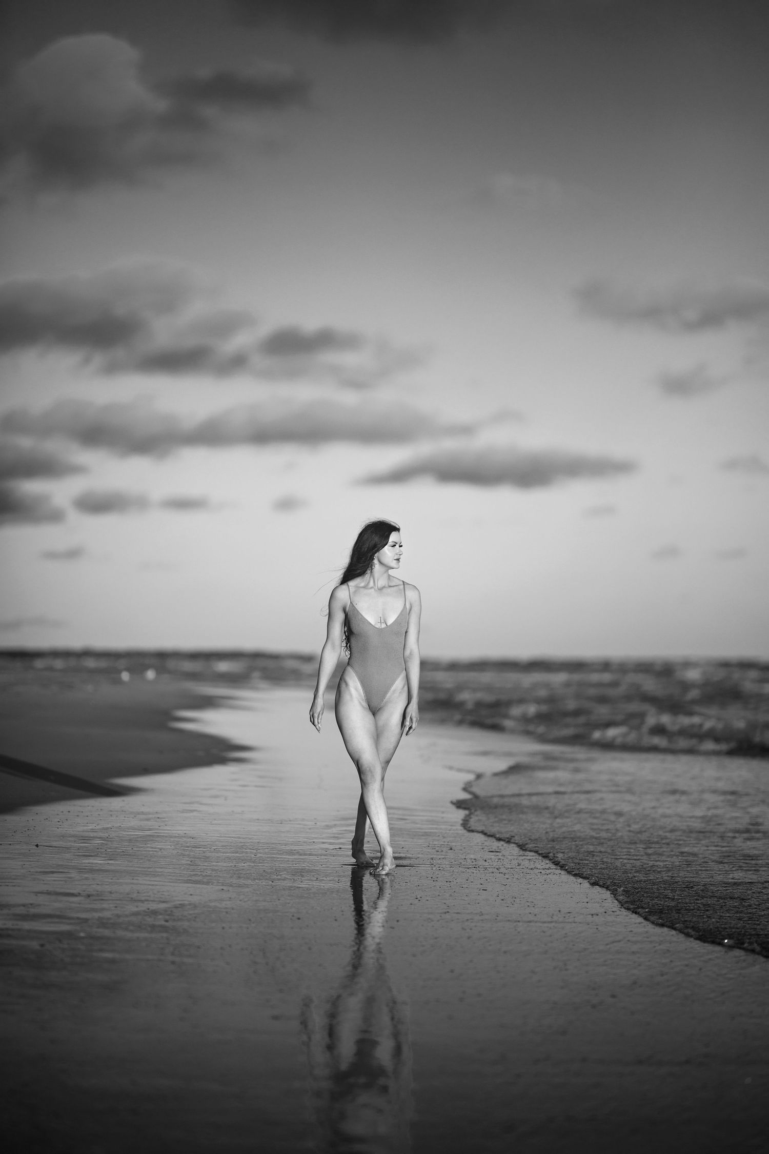 Black and white photo of a woman in a swimsuit walking along a wet beach shoreline at sunset.