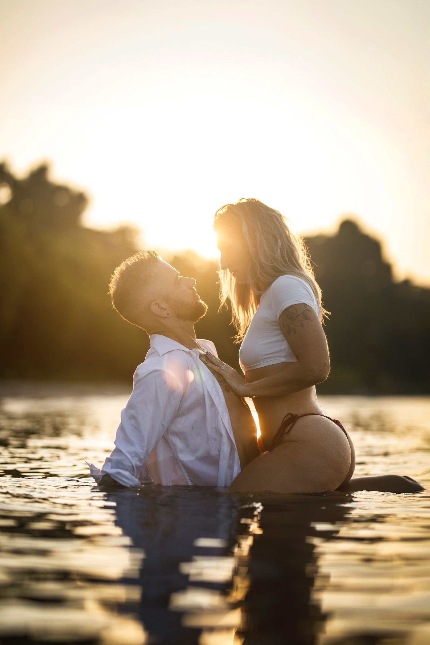 Romantic couple embracing in a sunlit lake at golden hour, woman in bikini sitting on mans lap in water.