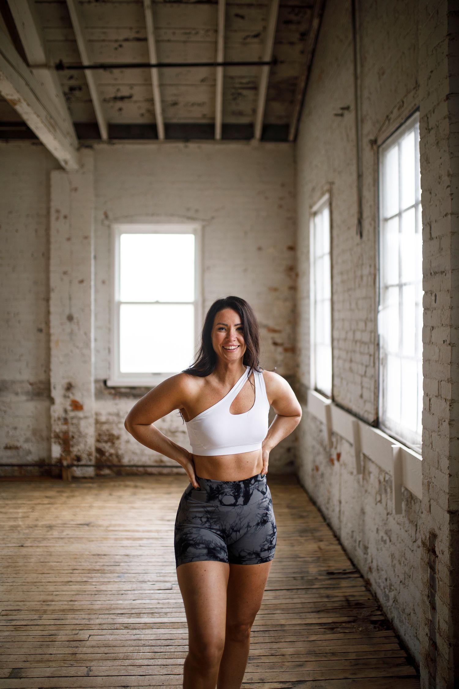 Woman in white sports bra and camo shorts posing confidently in a rustic brick studio with hardwood floors.