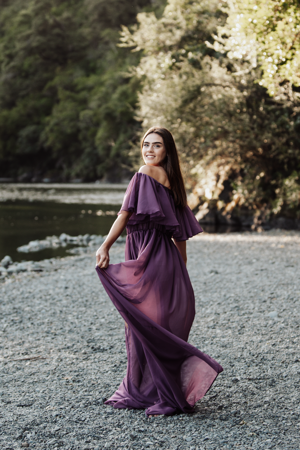 A flowing purple maxi dress with ruffled neckline catches the wind on a rocky beach at sunset.