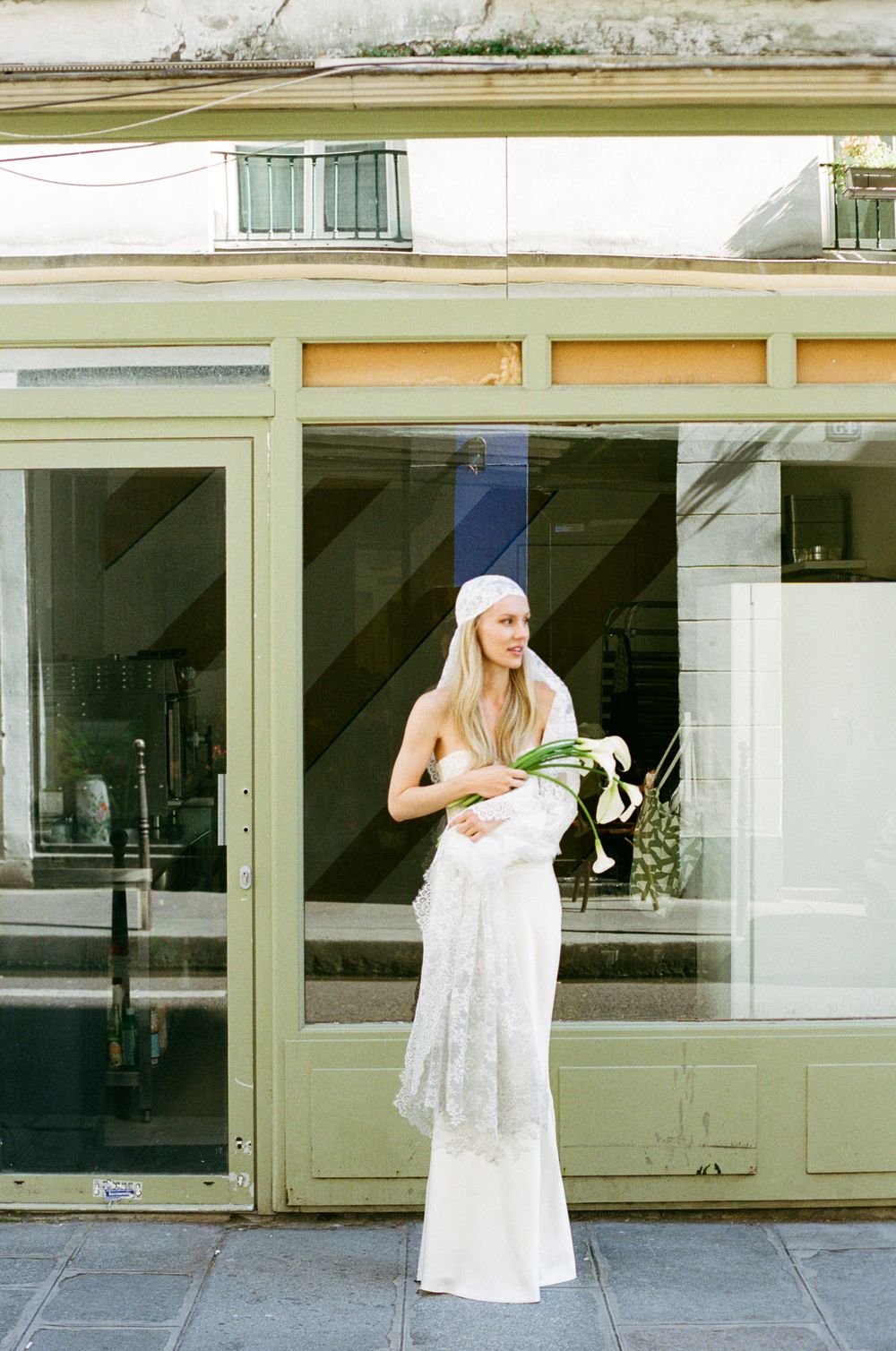 Bride in white lace gown and veil holding calla lilies stands before a green-framed storefront.