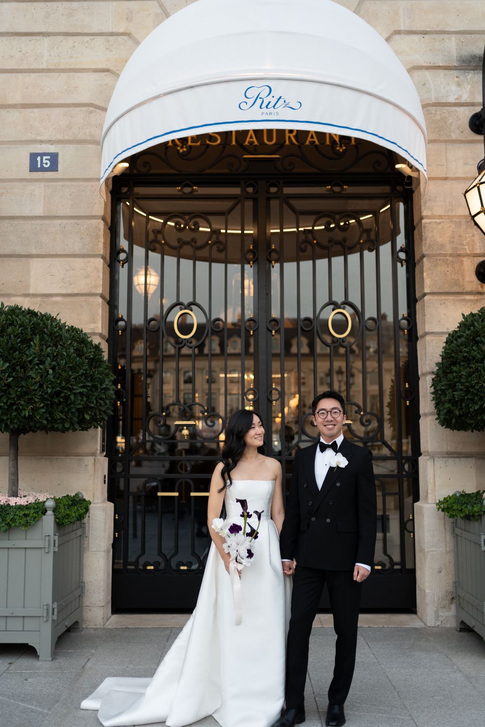 A bride and groom pose for wedding photos in front of an elegant restaurant entrance with potted topiaries. Ritz paris wedding. Ritz paris.