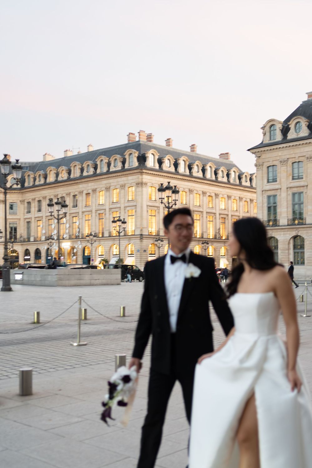 Bride and groom pose in front of illuminated Parisian building at dusk, bride in white gown, groom in black tuxedo.