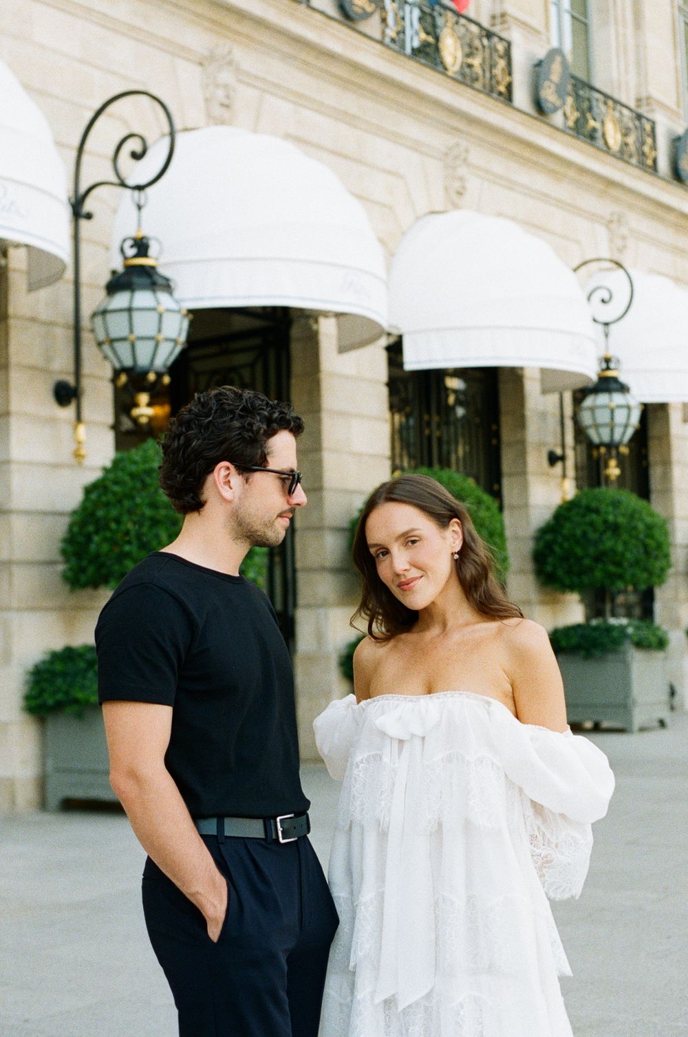 A couple poses outside a Parisian hotel, man in black outfit with sunglasses, woman in white off-shoulder dress.