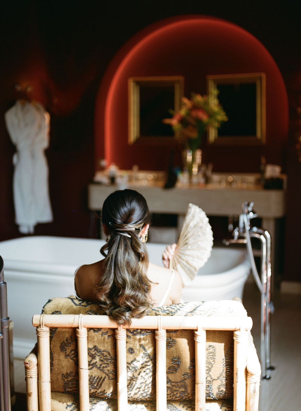 Bride with elegant half-up hairstyle sits in ornate chair holding fan, facing clawfoot bathtub in luxurious bridal suite.