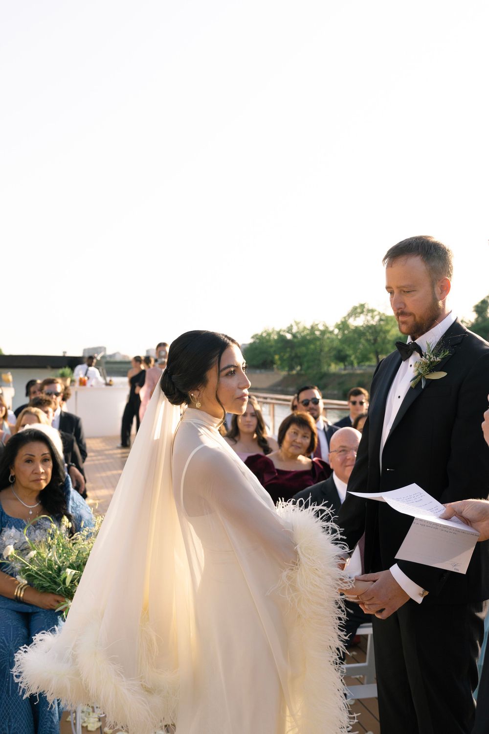 Bride in feather-trimmed tulle gown exchanges vows with groom in tuxedo at outdoor rooftop wedding ceremony.