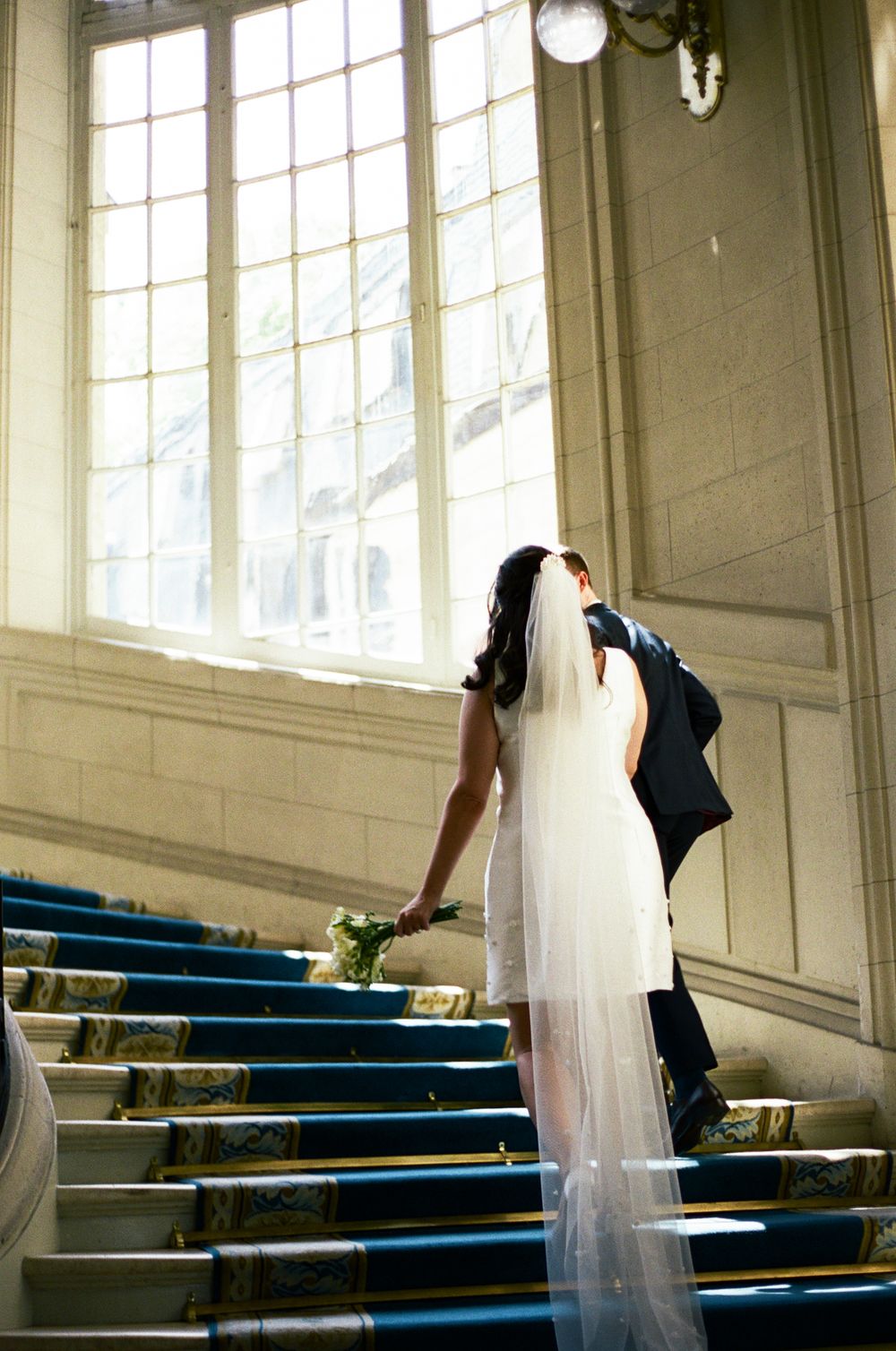 Bride and groom kissing on blue carpeted stairs near large window in elegant wedding venue.