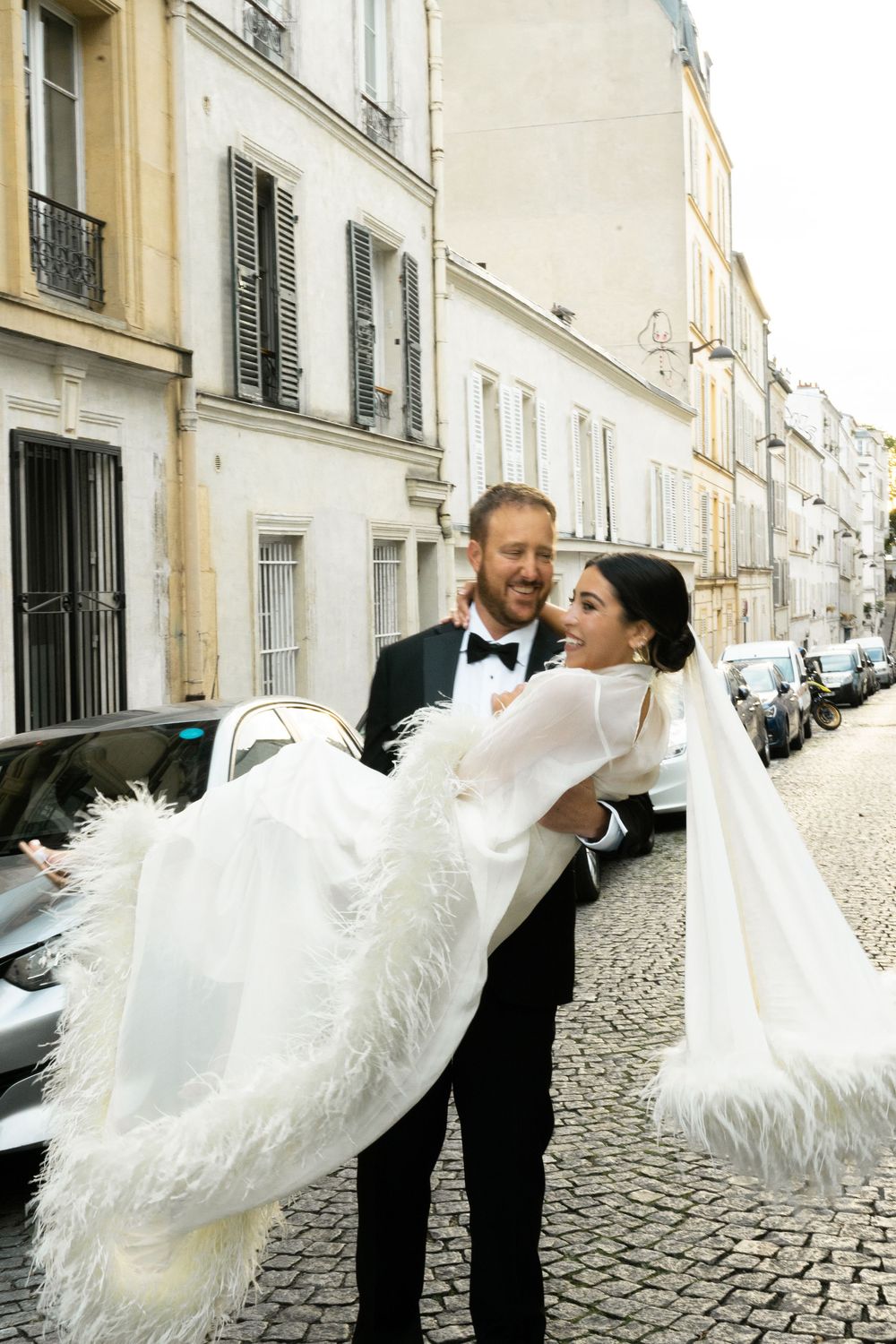 Groom in tuxedo carries bride in white feathered gown on cobblestone Paris street, both smiling joyfully.