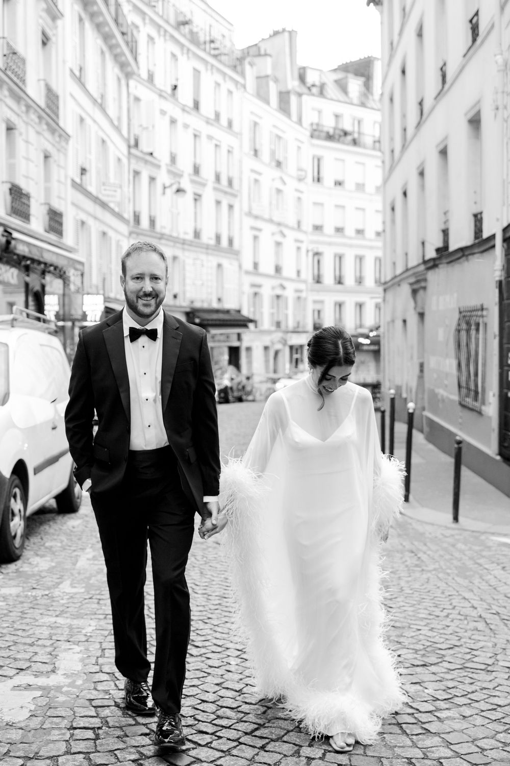A smiling groom in a tuxedo holds hands with his bride in a flowing white gown on a cobblestone Paris street.