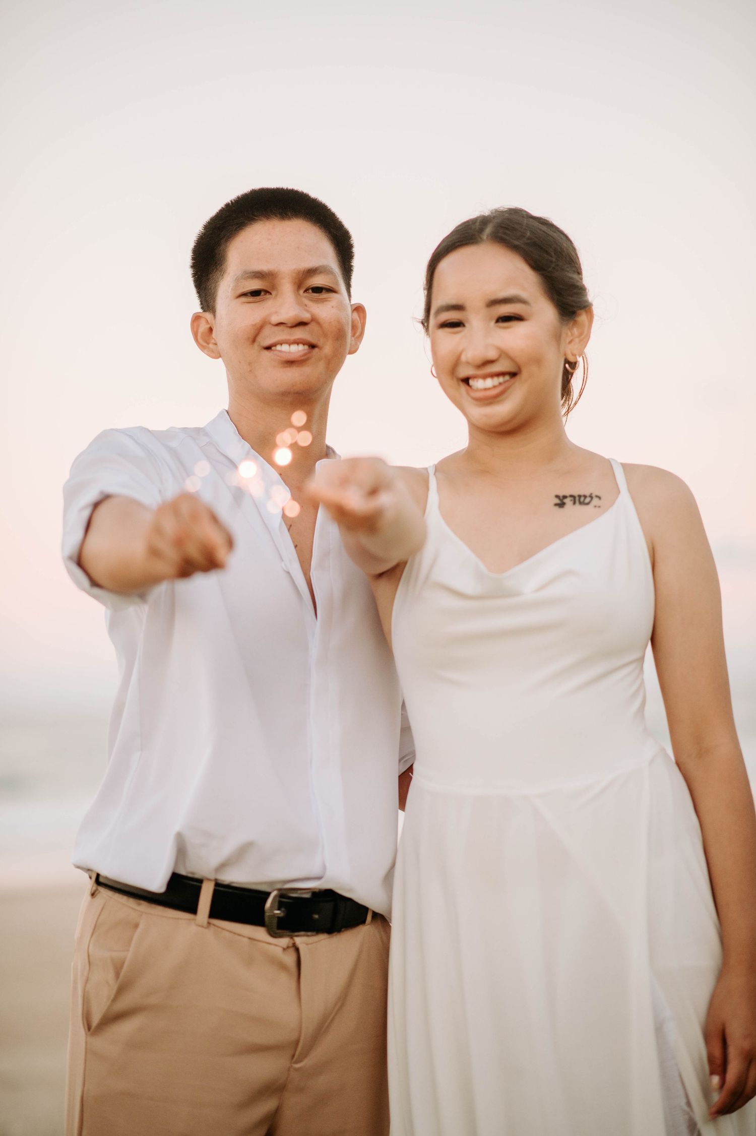A joyful couple in white and khaki outfits pose together against a sunset beach backdrop.