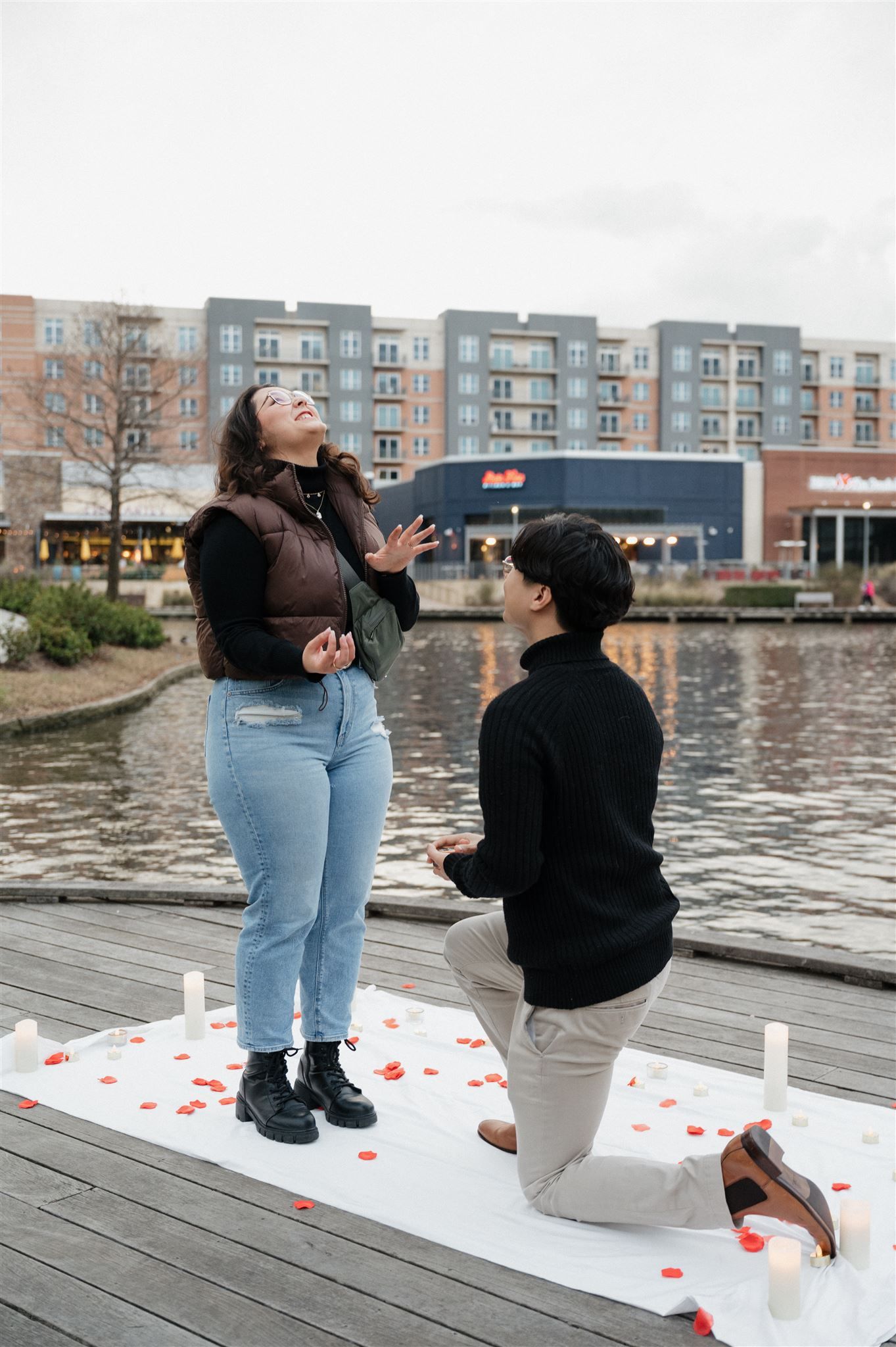 A romantic proposal moment on a waterfront dock decorated with rose petals and candles with apartments in the background.