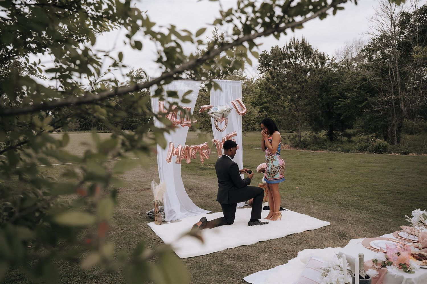 A romantic marriage proposal scene on a white blanket in a grassy outdoor setting with floral decorations.