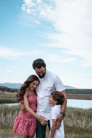 Family shares a tender moment together while standing in tall grass near a lake with mountains in the background.