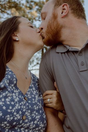 A romantic couple sharing a tender kiss outdoors, with her in a floral dress and him in a grey polo shirt.