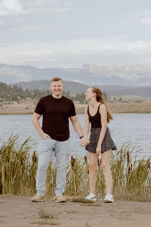 A couple stands together by a lake with mountains and tall grass in the background on a cloudy day.
