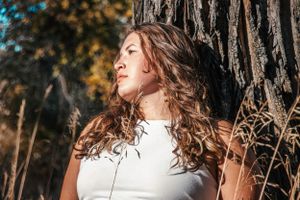 Sun-kissed portrait against tree bark with flowing curls and white top.