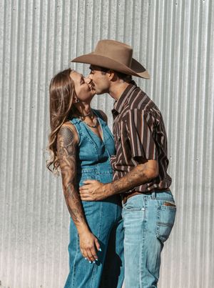 A couple in western style clothing share a romantic kiss against a metal wall backdrop.
