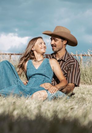 A romantic couple relaxes in a sunny field of tall grass, with the person wearing a cowboy hat and western attire.
