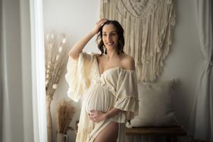 Giovane donna in gravidanza indossa un vestito romantico color panna. Con una mano si accarezza il pancione e con l'altra i capelli.