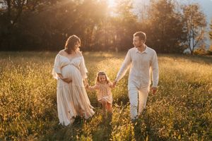 Famiglia composta da giovane mamma incinta, papà e figlia di 4 anni cammina mano nella mano in un campo abbracciata dalla luce del tramonto
