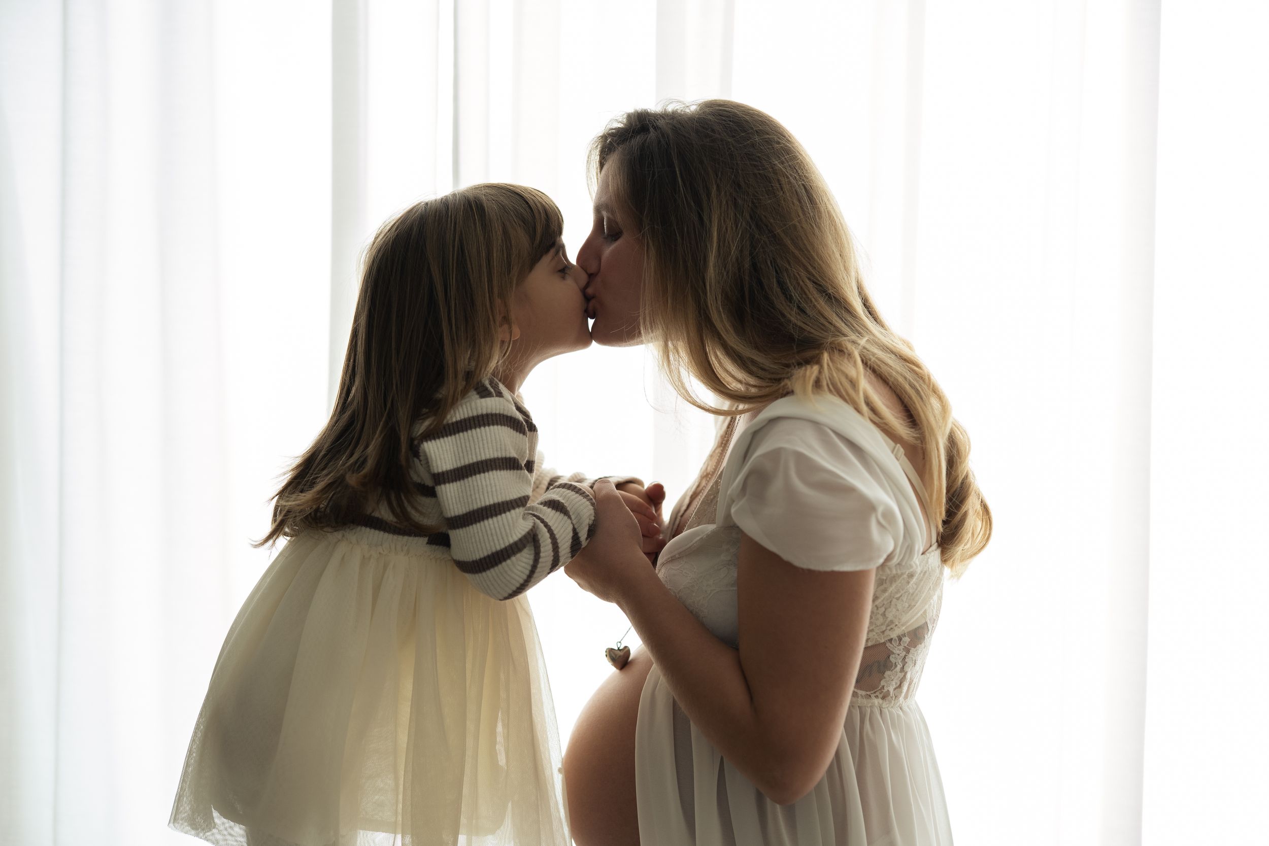 Mamma incinta e la sua bimba si danno un bacio affettuoso tenendosi le mani davanti a una tenda bianca che le illumina in controluce