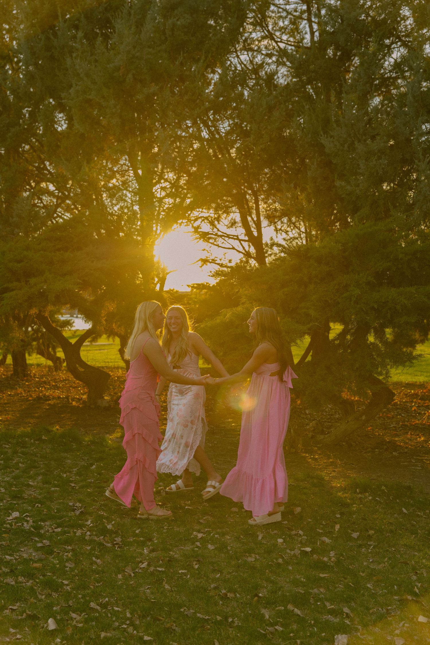 Women in pink and white dresses dance in a circle during golden hour in a lush garden setting.
