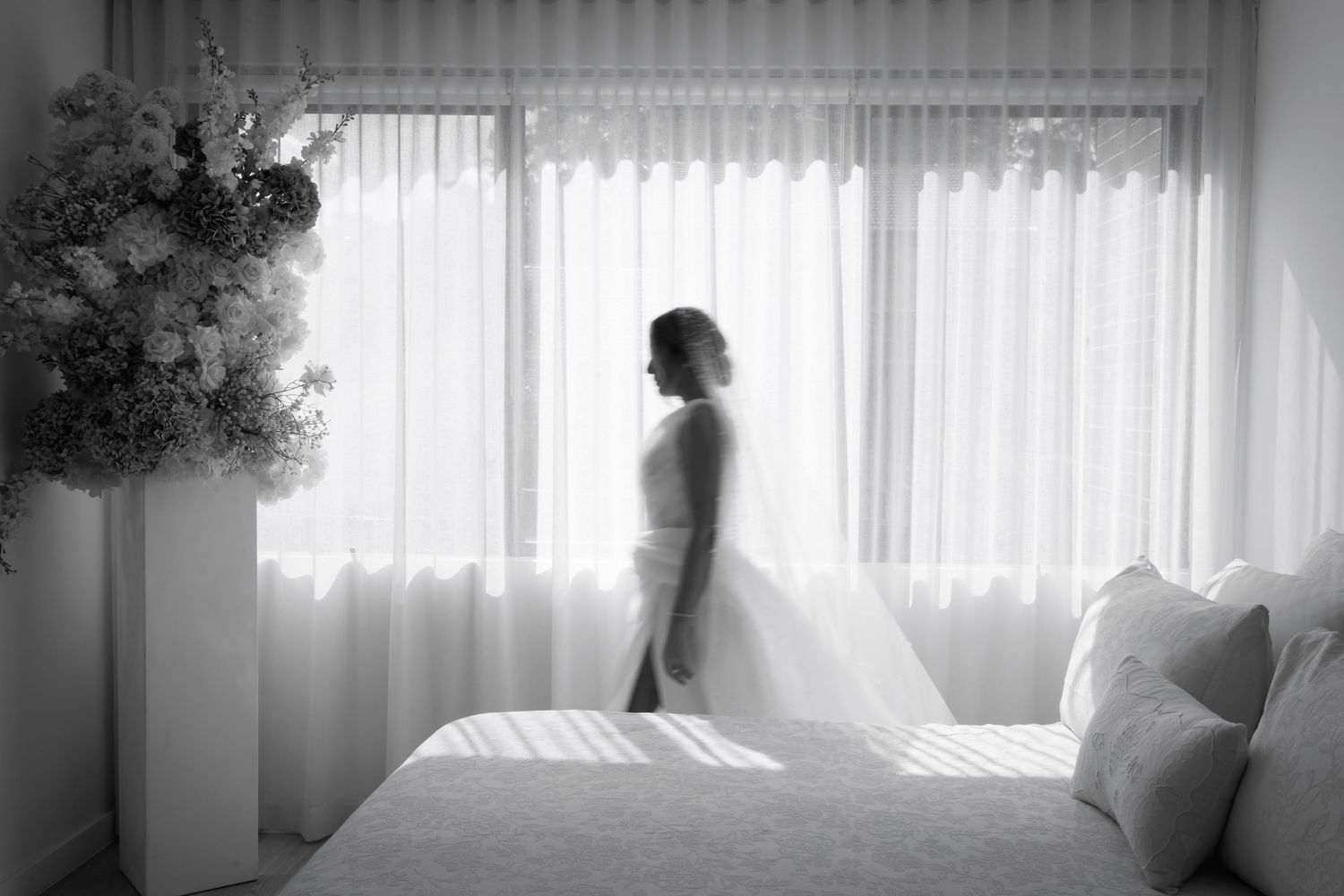 Bride in wedding dress standing by sheer curtained window in black and white bridal portrait.