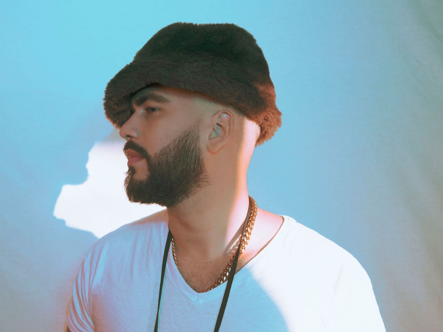 Bearded man wearing a flat cap and gold chain necklace posing against a light blue background.