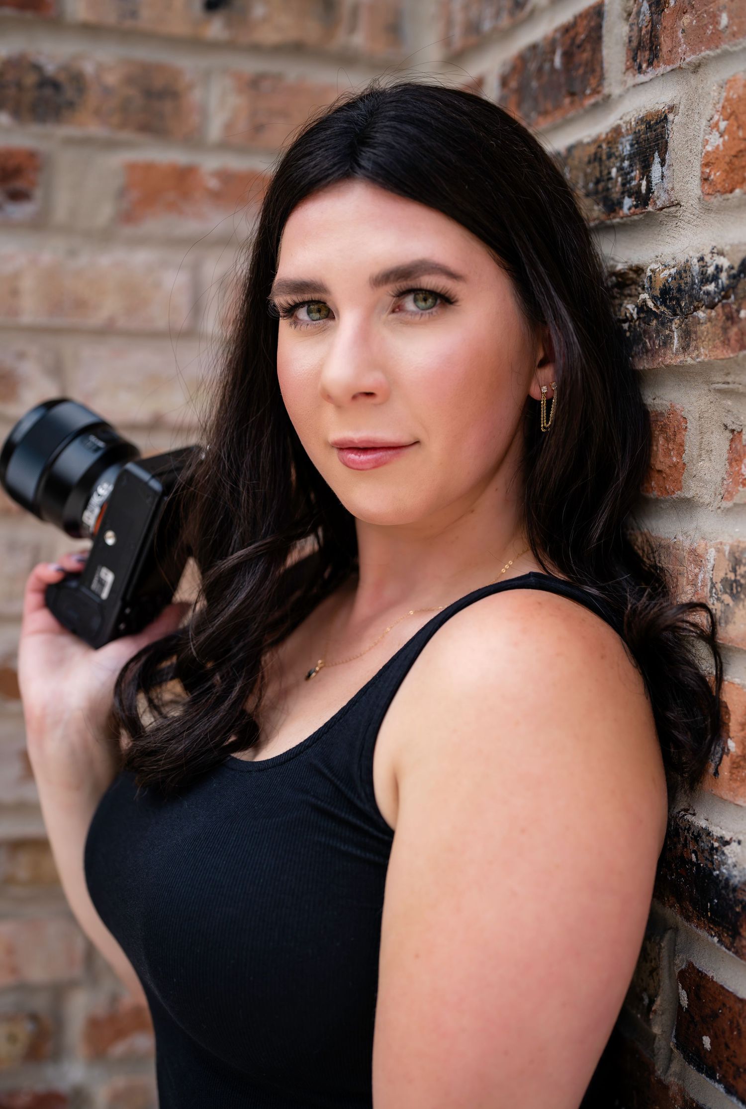 Photographer in black tank top poses with camera against rustic brick wall background.