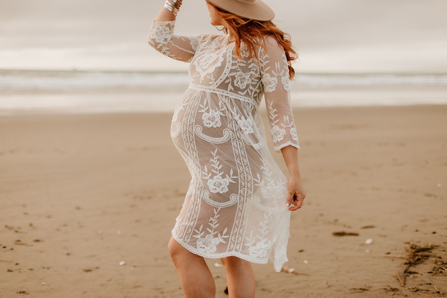 pregnant woman wearing a lace dress and a wide brimmed hat looking out into ocean while holding her flowing dress