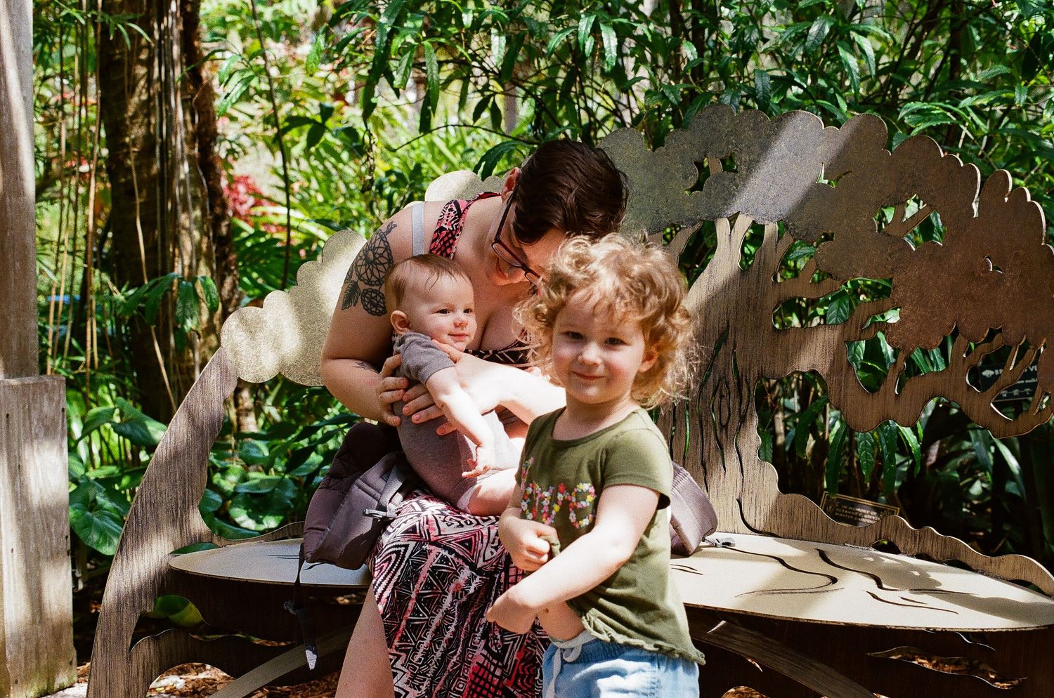 image of a young mother sitting on a bench with her infant son on her lap and her toddler daughter standing in front