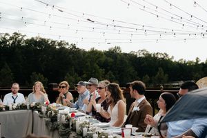 Guests seated at a long outdoor wedding reception table with string lights overhead and lush greenery in the background.