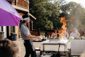 Chef grilling with open flames at outdoor BBQ event under purple umbrella with guests dining in background.