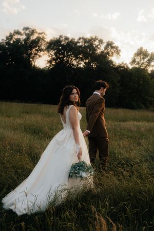 Bride in white gown holds bouquet, glancing back at groom in brown suit, standing in a lush green field at sunset.