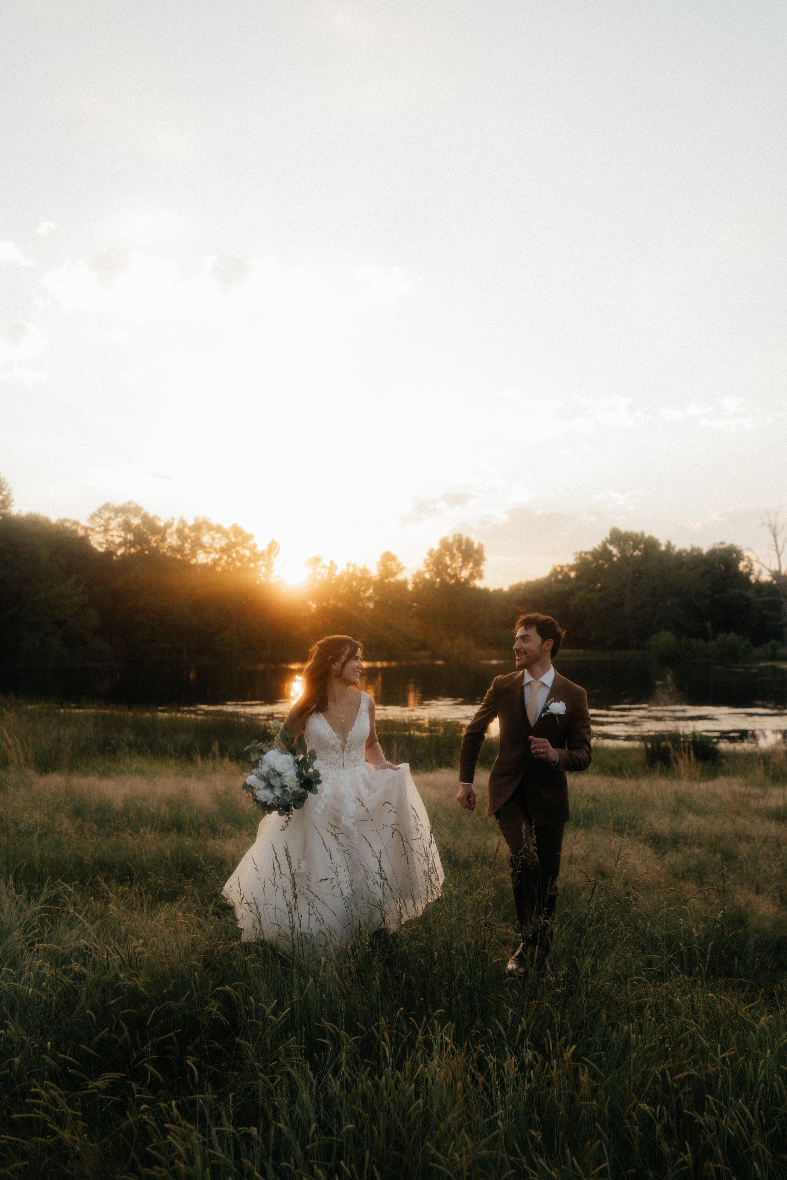 Bride and groom walk hand in hand through a sunlit meadow by a lake at golden hour sunset.