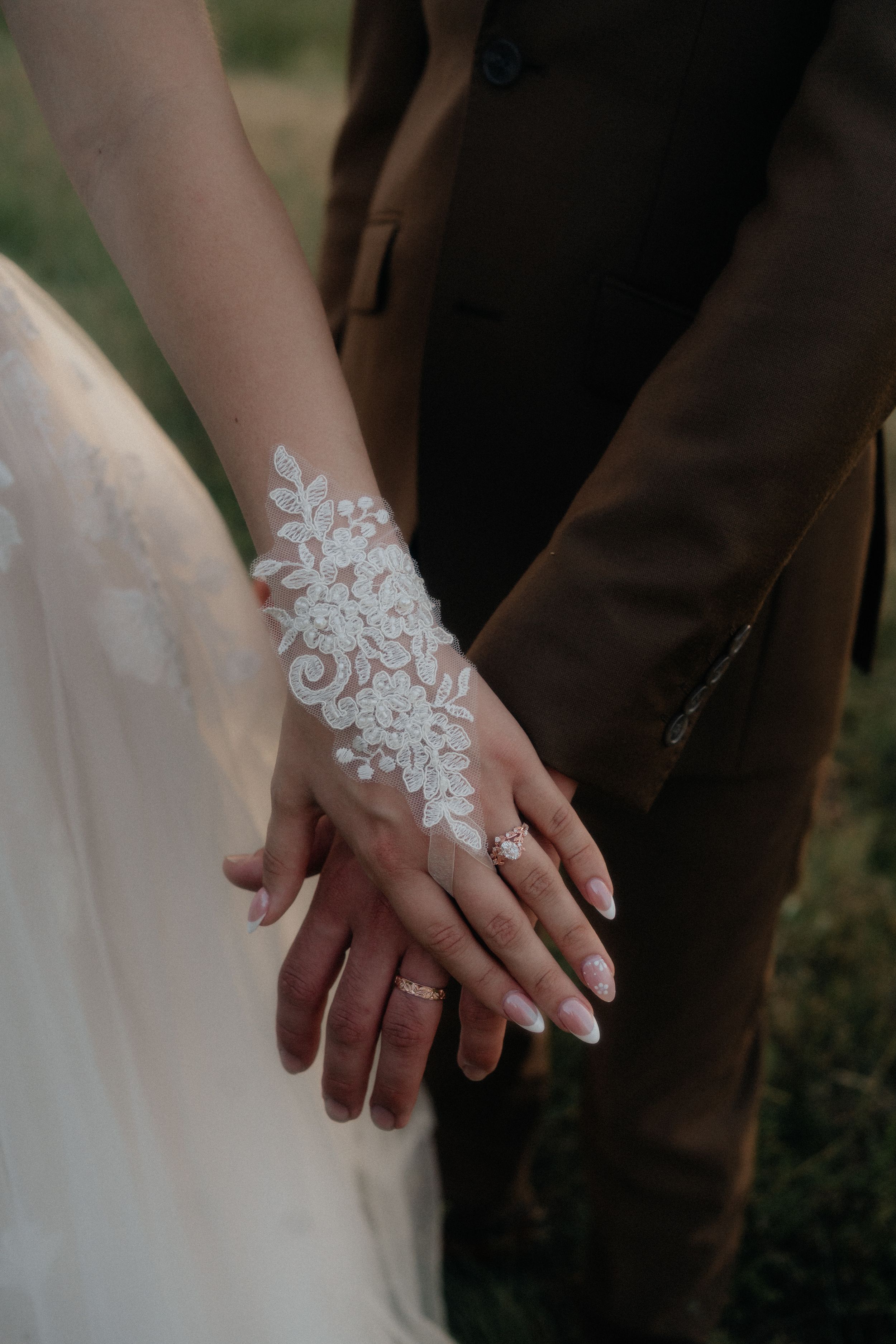 Bride and groom holding hands, showcasing lace glove and diamond ring, with groom in brown suit at wedding.
