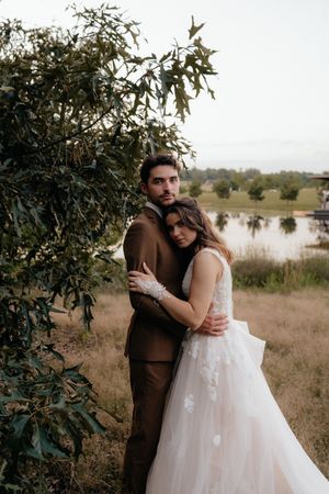 Bride and groom embracing under green foliage near a lake, bride in lace tulle gown, groom in brown suit.