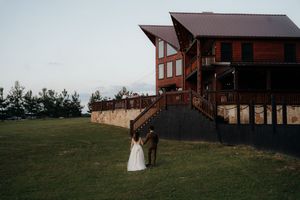 Bride and groom share a romantic moment on a green lawn beside a rustic wooden wedding venue at dusk.