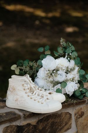 White embroidered Converse sneakers beside a bridal bouquet of white roses and eucalyptus on a stone surface.