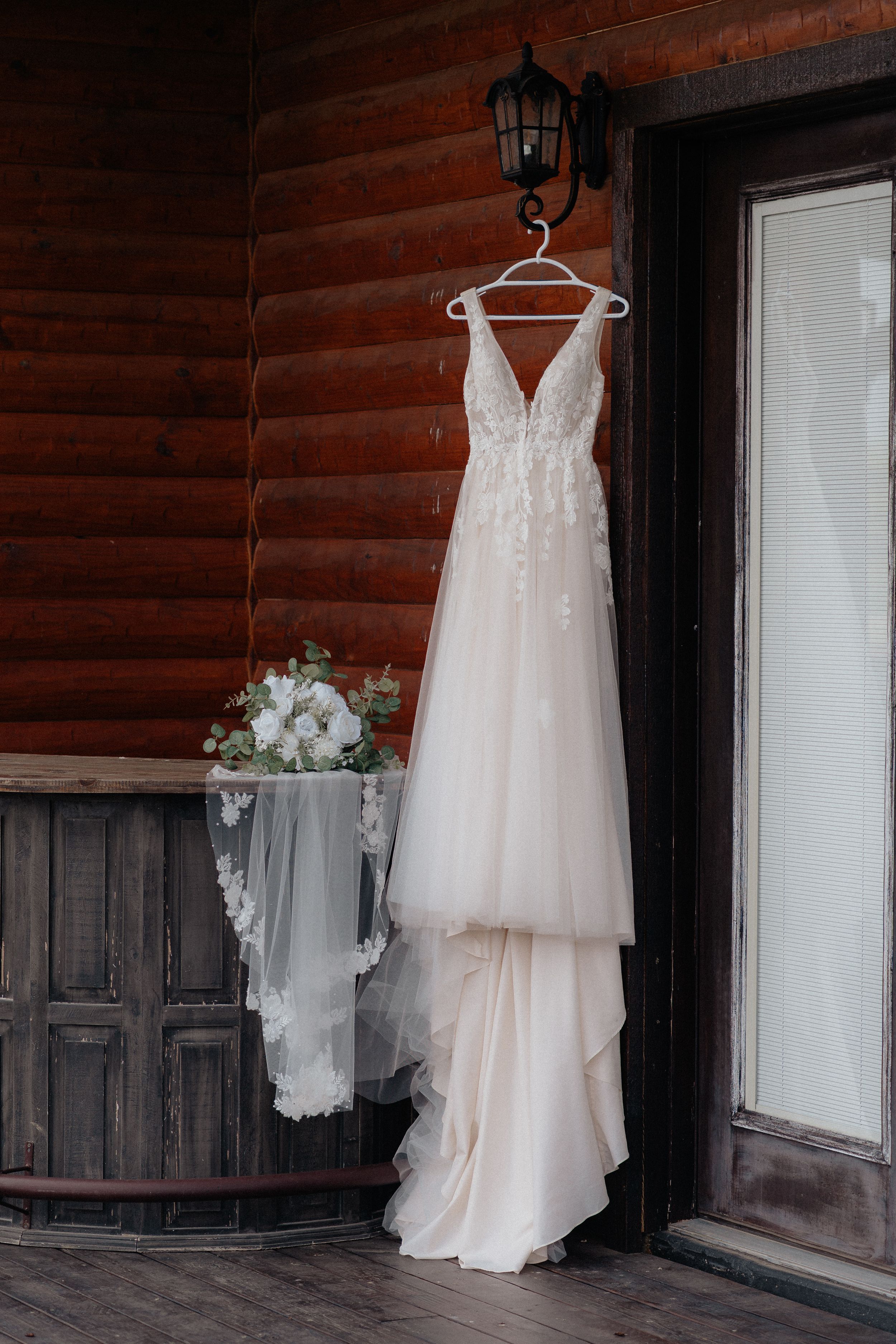 Elegant blush A-line wedding dress with lace bodice hanging on wooden cabin wall beside bridal bouquet.
