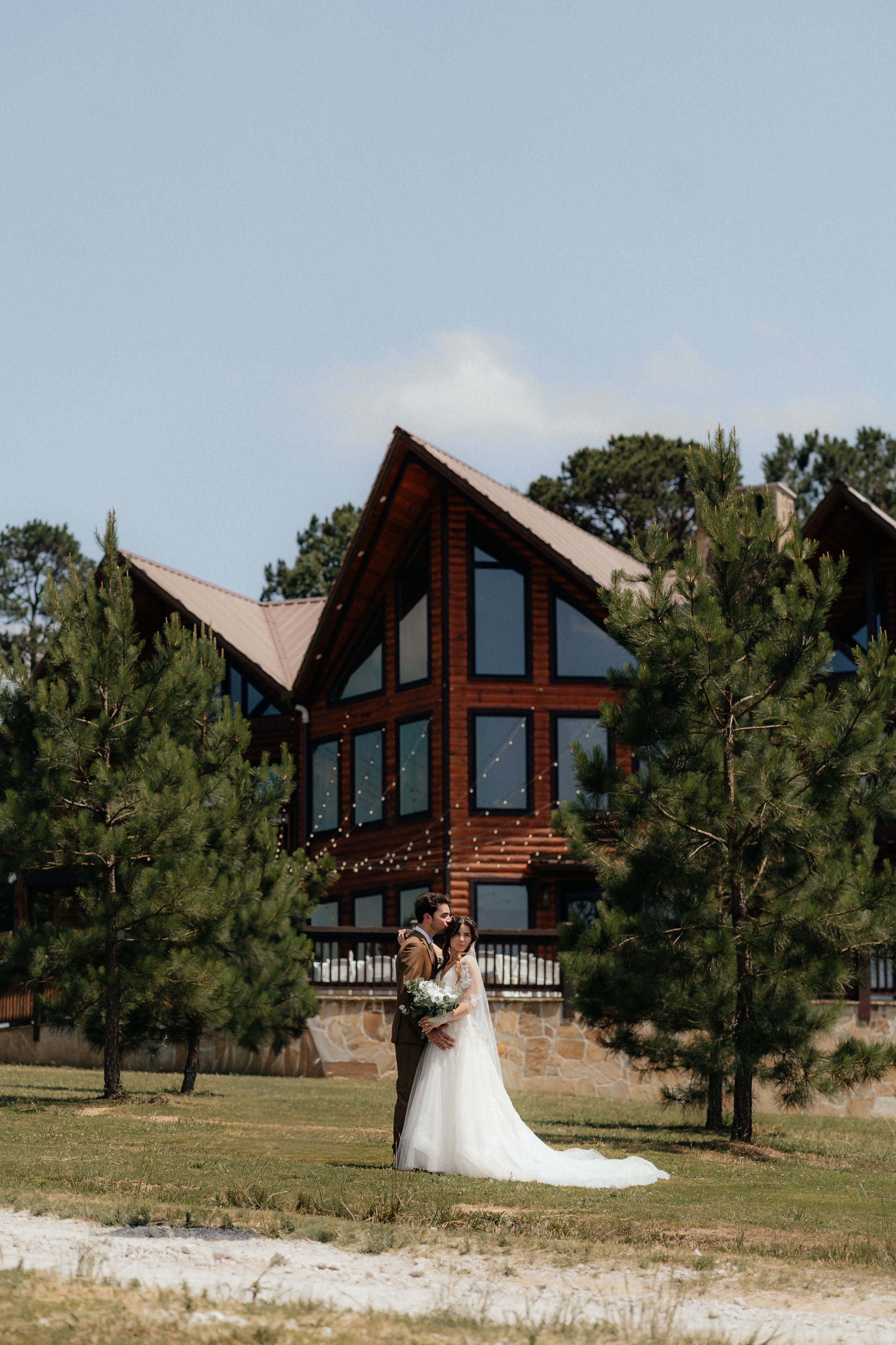 Bride and groom embrace in front of a rustic log cabin surrounded by tall pine trees on a sunny wedding day.