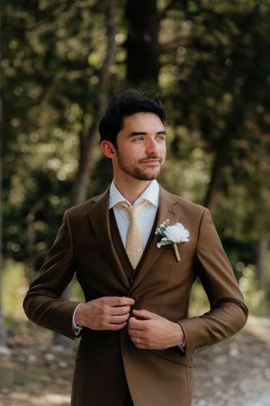 Groom in brown three-piece suit with white boutonniere and gold tie, adjusting jacket outdoors.