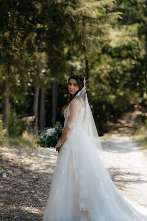 Bride in lace wedding gown and veil holds bouquet on a forest path.
