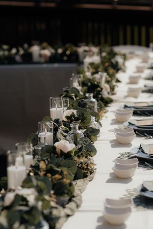 Wedding reception table with eucalyptus garland, white pillar candles, white bowls, and dark napkins.