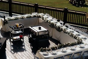 Elegant outdoor wedding reception setup on a wooden deck with white tablecloths, floral garlands, and white folding chairs.