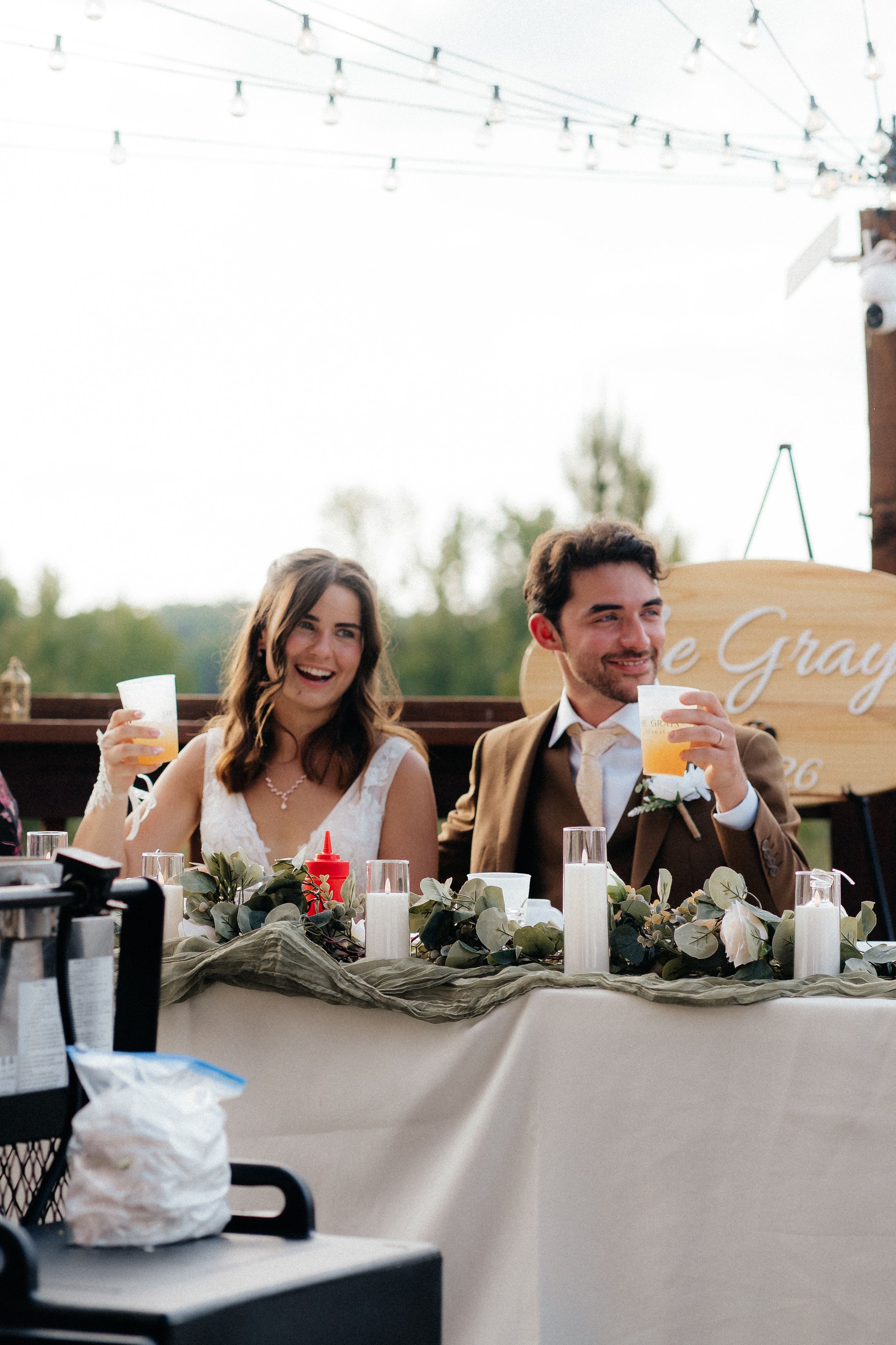 Bride and groom laugh while raising glasses at outdoor wedding reception table.