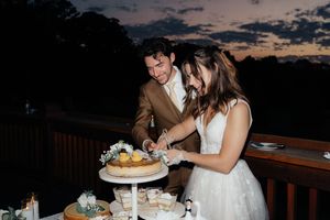 Bride and groom cutting wedding cake at sunset on outdoor deck, romantic twilight sky backdrop.