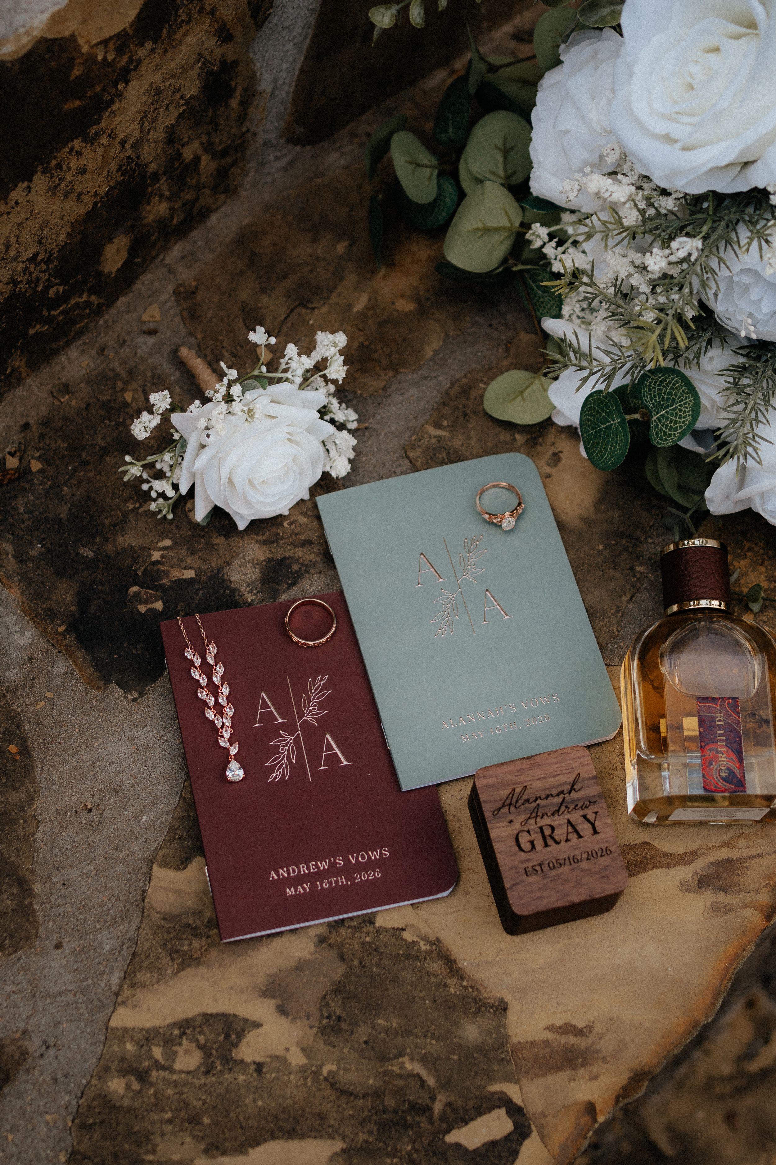 Wedding flat lay with vow books, rings, cologne, and white rose bouquet.